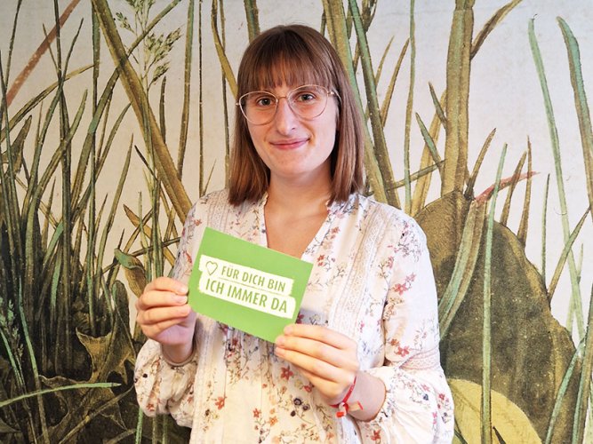 junge Frau mit Brille und Blumenkleid steht vor einer Wand mit Wiesenmotiv und hält Postkarte in der Hand "Für dich bin ich immer da"