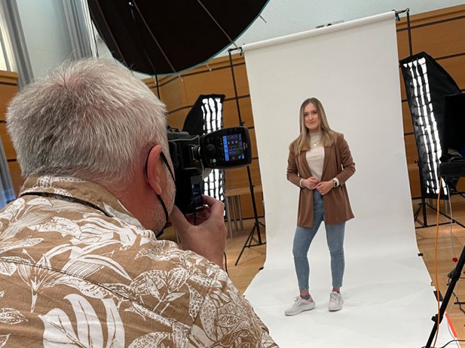 Fotograf im beigen Blumenhemd steht mit dem Rücken zur Kamera und macht ein Foto von einer jungen Frau in brauner Langjacke vor weißer Leinwand