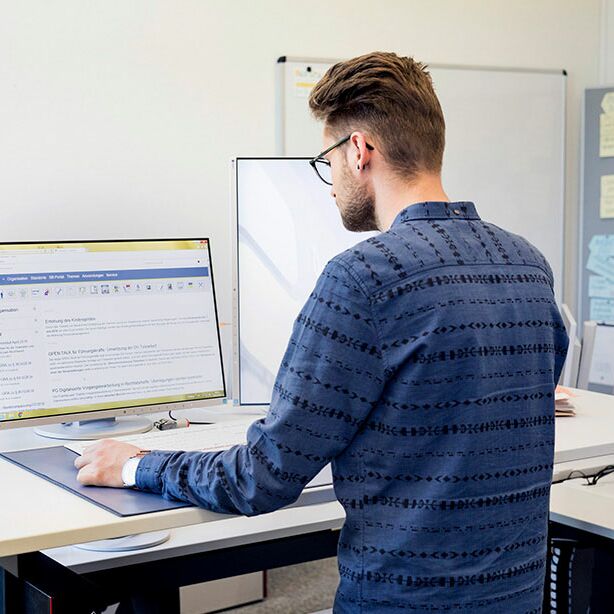 Ein Auszubildender steht an seinem ergonomischen Schreibtisch und arbeitet am PC.