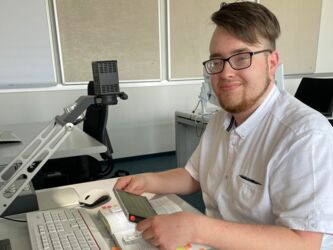 junger Mann mit Brille und weißem Hemd sitzt am Schreibtisch mit mobilem Lesegerät und elektronischer Lupe