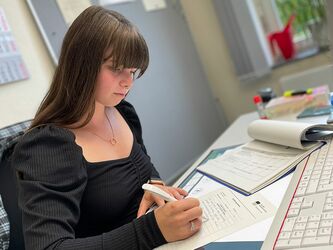 Frau mit schwarzem Shirt sitzt am Schreibtisch und füllt ein Dokument mit Kugelschreiber aus