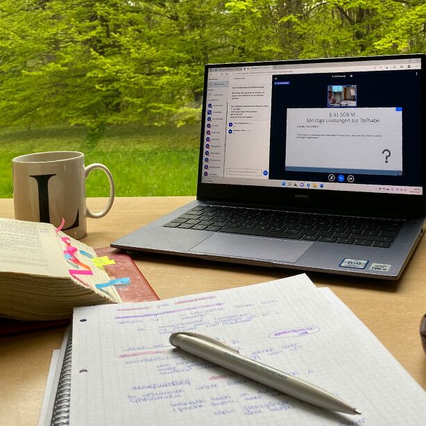 im Vordergrund Laptop, ausgeschlagenes Gesetzbuch und Schreibblock, im Hintergrund Blick ins Grüne