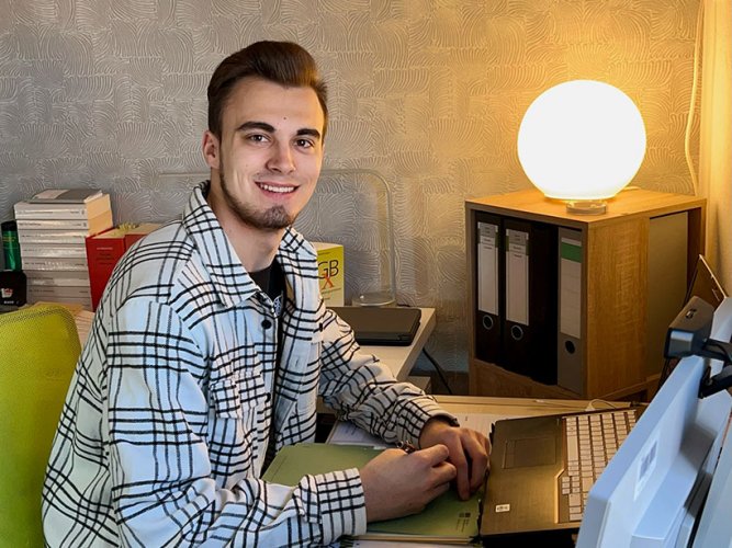 junger Mann im karierten Hemd sitzt am Schreibtisch mit Laptop und Stift in der Hand,hinter ihm stehen Ordner und Gesetzbücher