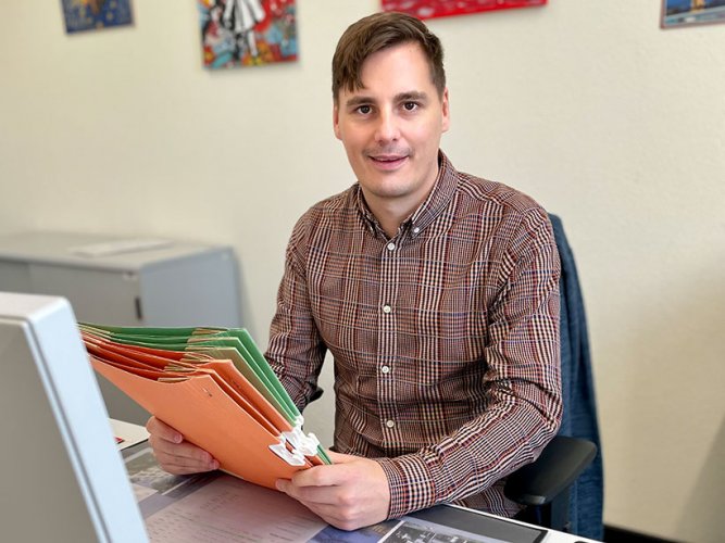 Mann mit kariertem Hemd und leeren Aktenhüllen in der Hand sitzt vor dem PC