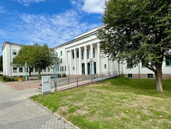 helles Hauptgebäude der Hochschule Meißen mit Säulen im Eingang, davor stehen Bäume und blühen Blumen
