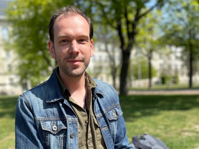 junger Mann in Jeansjacke sitzt vor Bäumen und dem Gebäude der Deutschen Rentenversicherung Mitteldeutschland in Leipzig