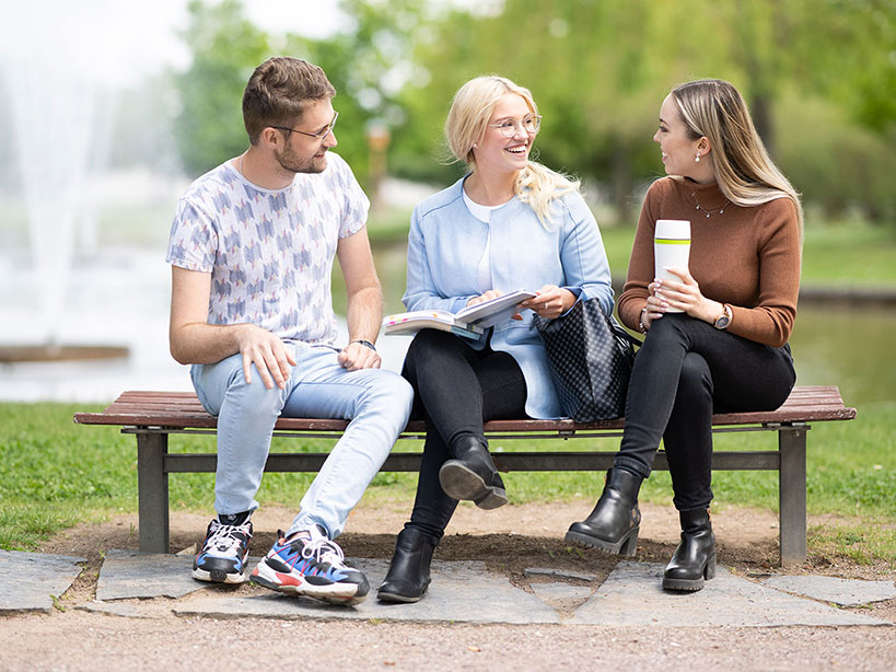 ein junger Mann und zwei junge Frauen sitzen auf einer Bank vor einem Springbrunnen und unterhalten sich