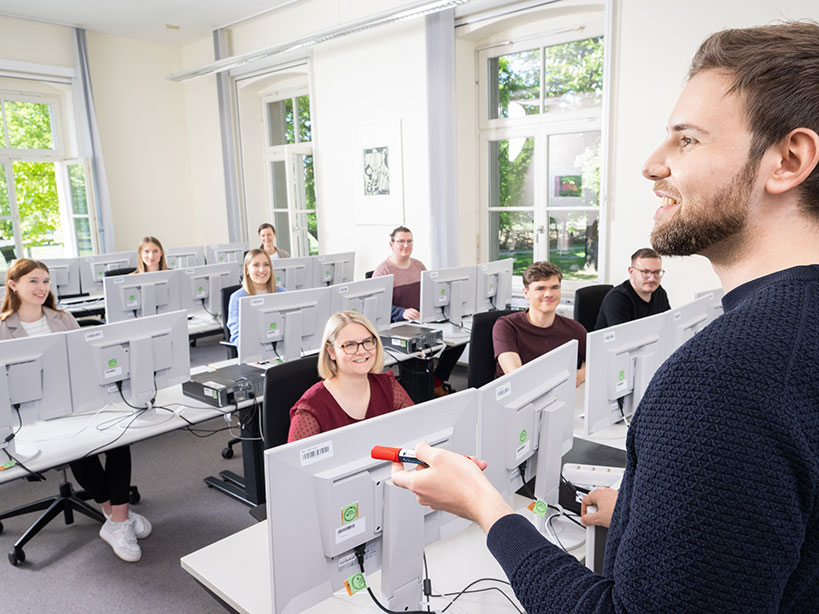 junge männliche Lehrkraft steht am rechten Bildrand und blickt in die Klasse, die am PC-Monitoren sitzt
