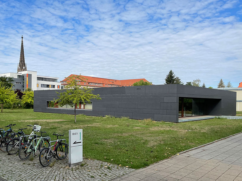 moderner anthrazitfarbener Flachbau der Bibliothek, umgeben von Wiese, im Hintergrund ein Kirchturm