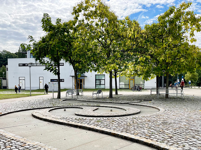 gepflasterter Brunnen in Form eines Paragrafen, umgeben von Bäumen und Bänken, im Hintergrund das Gebäude der Mensa
