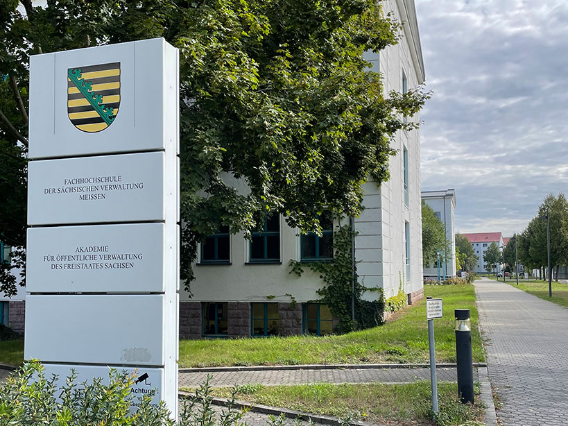 Wegweiser-Schild der Fachhochschule der sächsischen Verwaltung Meißen mit sächsischem Wappen, dahinter beginnen die Gebäude der Hochschule